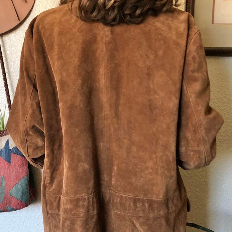 May include: A brown suede jacket with short sleeves and two front pockets. The jacket has a collar and a button closure. The suede is a warm, earthy tone, and the jacket appears to be vintage.