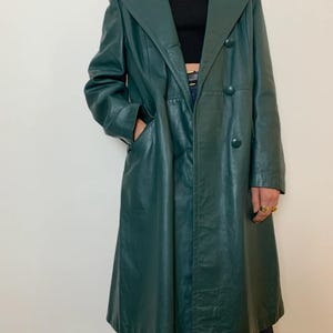 May include: A long, emerald green leather coat with a double-breasted design. The coat features a notched collar, two front pockets, and large buttons. The coat is knee-length and has a classic, tailored silhouette.