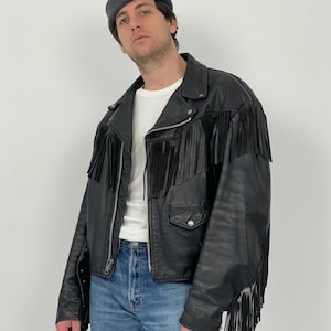 Puede incluir: Chaqueta de cuero negra con flecos en el pecho, los hombros y las mangas. La chaqueta tiene cierre de cremallera, dos bolsillos delanteros y un diseño clásico de chaqueta motera. La persona lleva una camiseta blanca y vaqueros azules.