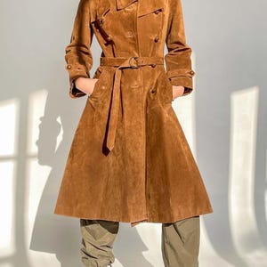 May include: A brown suede trench coat with a double-breasted design, a belted waist, and buttoned cuffs. The coat has a flared skirt and is worn with olive green pants and black high-heeled boots.