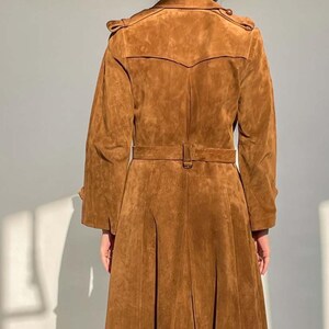 May include: A brown suede trench coat with a belted waist and pleated skirt. The coat features a classic trench coat design with shoulder epaulets and a notched collar. The long coat is a warm, earthy tone.