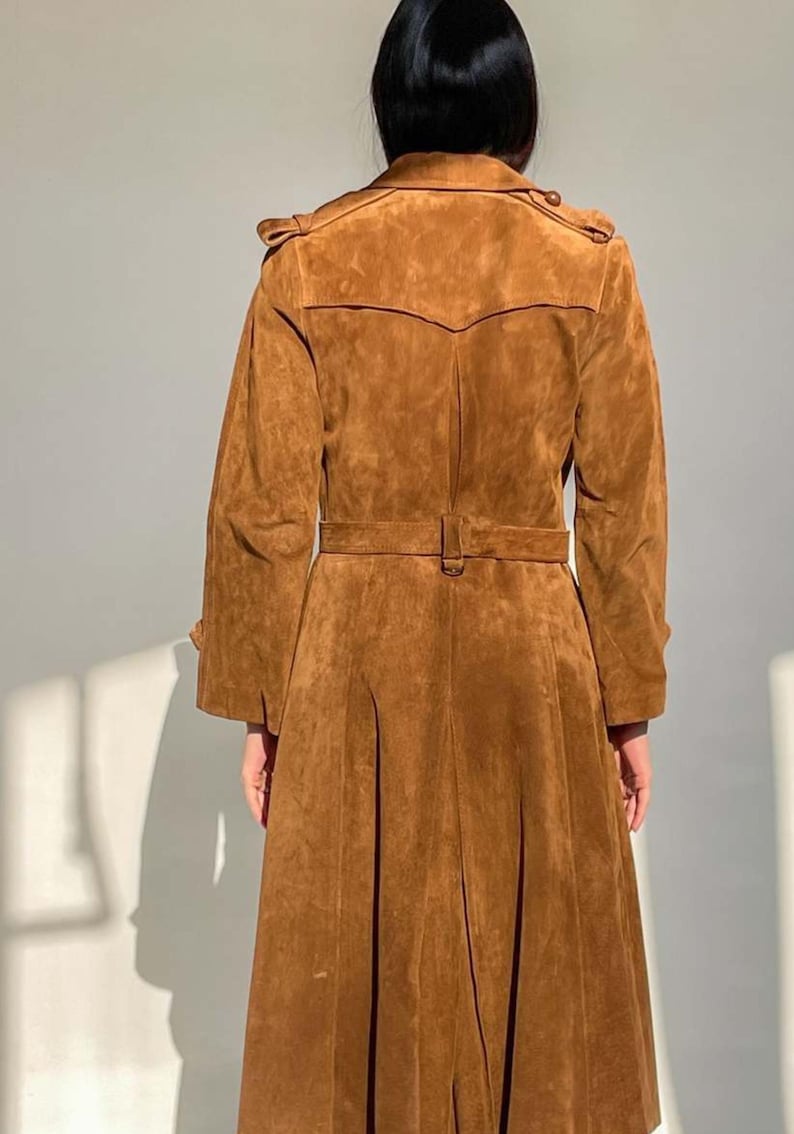 May include: A brown suede trench coat with a belted waist and pleated skirt. The coat features a classic trench coat design with shoulder epaulets and a notched collar. The long coat is a warm, earthy tone.
