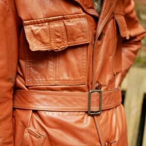 May include: A vintage, russet-colored leather jacket with a belted waist and multiple pockets. The jacket features a classic collar and button closures. The leather appears worn, adding to its vintage appeal.
