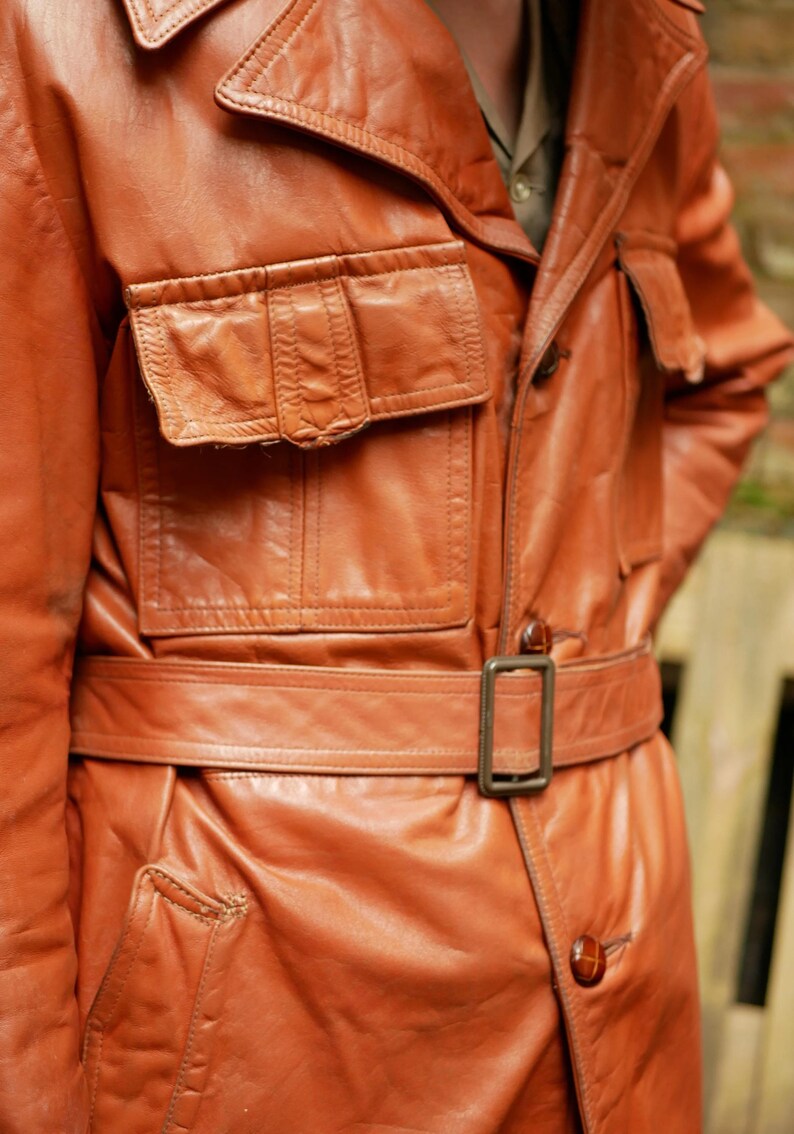 May include: A vintage, russet-colored leather jacket with a belted waist and multiple pockets. The jacket features a classic collar and button closures. The leather appears worn, adding to its vintage appeal.