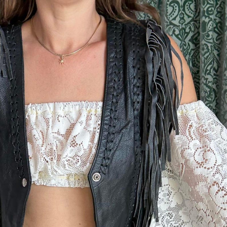 May include: A black leather vest with fringe detail and a white lace tube top. The vest has braided accents and a button closure. A gold necklace with a starburst pendant is also visible.