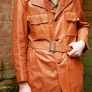May include: A vintage, tan leather jacket with a belted waist and multiple pockets. The jacket features a classic collar, button closures, and epaulettes. The leather appears worn, giving it a retro aesthetic. The jacket is a medium length, and the belt is secured with a rectangular buckle.