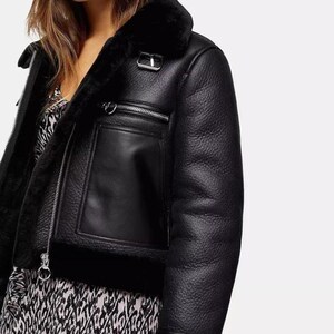 May include: Black faux leather jacket with a shearling lining and collar. The jacket features a zippered front, a chest pocket, and a buckle detail at the collar. The jacket is a cropped style.