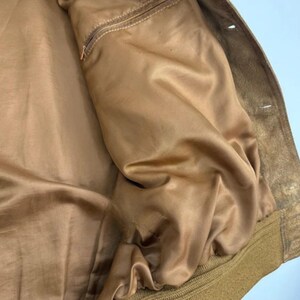 May include: Close-up of a tan suede jacket interior. The jacket features a smooth, satin-like lining in a matching tan color. A small zippered pocket is visible on the left side. The jacket's exterior is a textured suede.