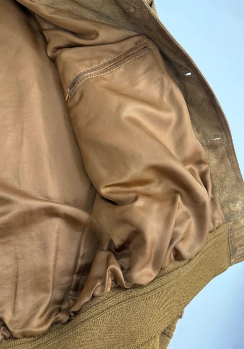 May include: Close-up of a tan suede jacket interior. The jacket features a smooth, satin-like lining in a matching tan color. A small zippered pocket is visible on the left side. The jacket's exterior is a textured suede.