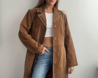 May include: A brown suede coat with a button-down front and white stitching details. The coat has a classic lapel collar and a tailored fit. The coat is knee-length and is paired with blue jeans.