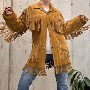 May include: A tan suede jacket with long fringe detailing. The jacket has a button-up front, two front pockets, and a collar. The fringe is along the sleeves, chest, and hem. The model is wearing blue jeans.
