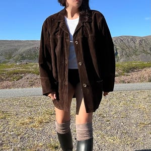 May include: A dark brown suede jacket with a button-up front, worn over a white top. The jacket is paired with black boots and knee-high socks. The background features a clear blue sky and a gravel path.