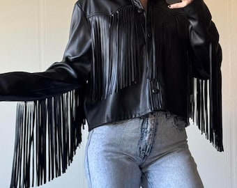 Puede incluir: Chaqueta negra de cuero sintético con flecos largos en las mangas y el pecho. La chaqueta tiene cuello y cierre de botones. Las mangas son largas y la chaqueta es corta.