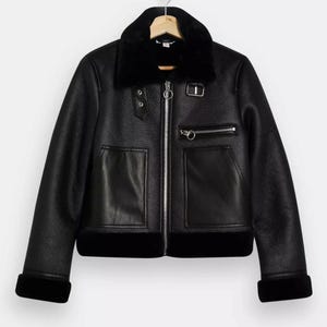 May include: Black faux leather jacket with a black faux fur collar, cuffs, and hem. The jacket features a silver zipper, two front pockets, and a small zippered chest pocket. The jacket is hanging on a wooden hanger.