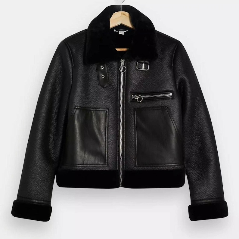May include: Black faux leather jacket with a black faux fur collar, cuffs, and hem. The jacket features a silver zipper, two front pockets, and a small zippered chest pocket. The jacket is hanging on a wooden hanger.
