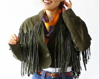 May include: Olive green suede fringe jacket with a brown leather belt and blue jeans. A colorful scarf is worn around the neck. The jacket has long fringe detailing on the sleeves and front.