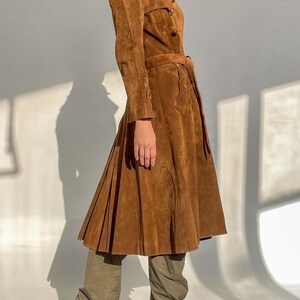 May include: A brown suede trench coat with a belted waist and pleated skirt. The coat features a double-breasted closure and long sleeves. The model is wearing olive green pants and black pointed-toe high heels.