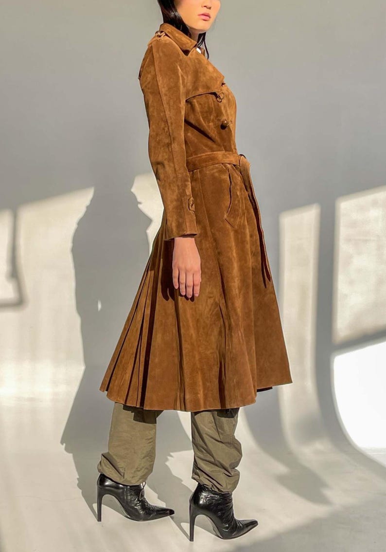 May include: A brown suede trench coat with a belted waist and pleated skirt. The coat features a double-breasted closure and long sleeves. The model is wearing olive green pants and black pointed-toe high heels.