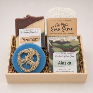 May include: A wooden gift box containing two rectangular bars of handmade artisan soap labeled Piedmont and Alaska, a blue circular soap with a loofah center, and a white textured bio-plastic soap saver with a paper band label.