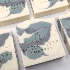 May include: Close-up of several rectangular bars of soap. The soap has a creamy white base with a marbled design in shades of gray and teal. The text "Little River Soapery" is embossed on the top of each bar.