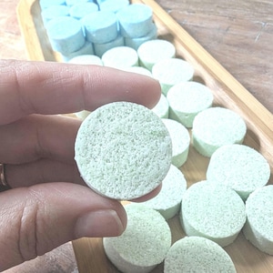May include: A hand holds a light green, round tablet with a textured surface. Other similar tablets are in a wooden tray, some light green and some light blue. The tablets appear to be bath bombs or similar products.