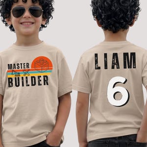May include: A beige short-sleeve t-shirt featuring a front graphic with the text MASTER BUILDER, a construction vehicle, tools, and a building block inside a sunset circle. The back displays the name LIAM and the number 6 in black and white.