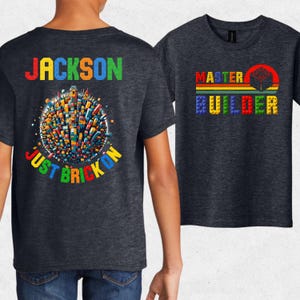 May include: A dark heather gray t-shirt featuring a graphic print. The back displays the name JACKSON above a spherical city made of colorful building blocks and the text JUST BRICK ON. The front shows the text MASTER BUILDER in block letters.