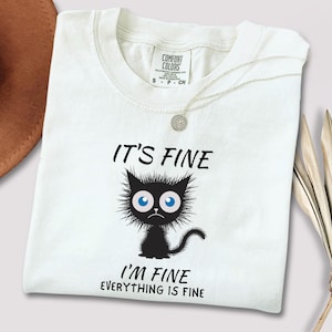 May include: White t-shirt with the phrase "IT'S FINE I'M FINE EVERYTHING IS FINE" and a cartoon black cat with blue eyes. The shirt has a crew neck and a small tag at the neckline.