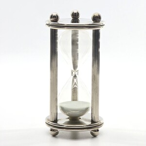 May include: A silver-toned hourglass featuring a clear glass bulb filled with white sand. The frame consists of three fluted vertical pillars connecting a circular top and base, each accented with three small spherical finials at the edges.