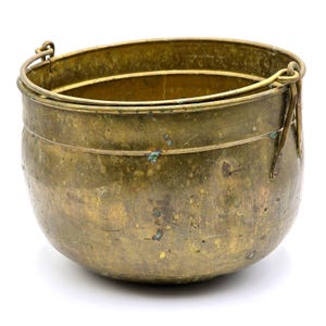 May include: A round brass pot with a weathered, tarnished finish and visible green oxidation spots. The vessel features a wide, open top and a sturdy, hinged metal handle attached to two side loops, allowing the handle to swing freely.