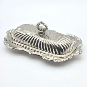 May include: A silver-colored rectangular butter dish with a removable lid. The lid features a fluted, ribbed design and a central decorative handle. The base has a scalloped, ornate edge with embossed detailing along the entire perimeter.