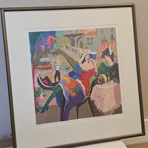 May include: Colorful framed print of two figures in Venice, Italy. One figure wears a red dress and blue hat, the other a green dress. A gondola and bridge are in the background. The print is in a gold frame.