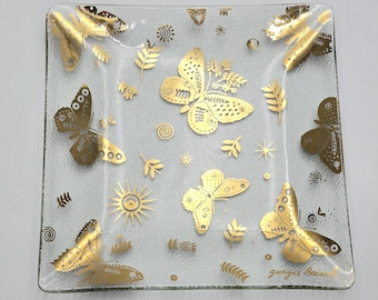 May include: A square clear glass tray featuring a gold butterfly pattern with various botanical elements, sun motifs, and small swirls. The signature Georges Briard is printed in gold in the bottom right corner of the textured glass surface.