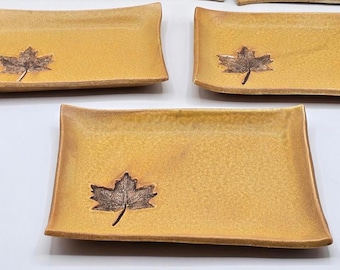 May include: Set of rectangular ceramic plates in a warm yellow hue, each adorned with a detailed brown leaf design. The plates have slightly curved edges and a textured surface, perfect for serving appetizers or small dishes.