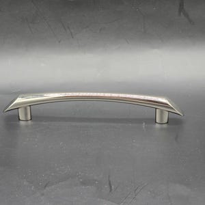 May include: A brushed nickel cabinet pull with a curved, arched design. The handle has a rectangular base and tapers to a point on each end. The overall length is approximately 6 inches.