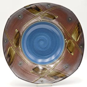 May include: A round ceramic bowl featuring a smooth, solid blue center with a visible spiral pattern. The wide rim is a deep reddish-brown color, decorated with dark brown brushstroke accents, thin intersecting lines, and small white dots.