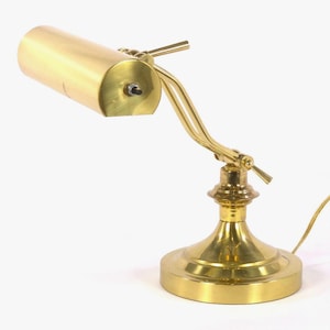 May include: A polished brass desk lamp featuring a cylindrical shade, an adjustable arm with a central hinge, and a circular base. The lamp includes a small black power switch located on the side of the shade and a thin gold power cord extending outward.