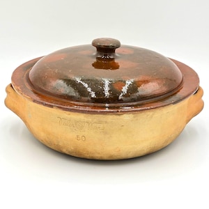 May include: A round, tan-colored earthenware casserole dish with two side handles and a matching dark brown glazed lid. The side of the dish features the embossed text Valor Ware and the number 50. The lid has a central knob handle.