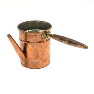 May include: A cylindrical copper pot with a metallic interior, a long angled spout, and a horizontal wooden handle attached with a brass fitting. The exterior surface shows a weathered patina with visible signs of oxidation and metallic texture.
