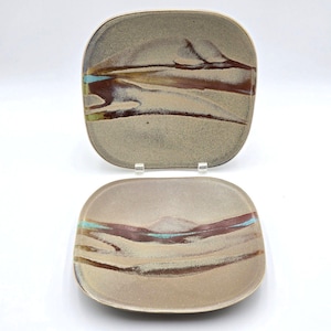 May include: Two square ceramic plates with rounded corners, featuring a speckled beige base glaze. Each plate displays horizontal, abstract brushstrokes in shades of dark brown and a vibrant turquoise accent line across the center.