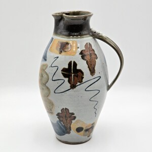 May include: A ceramic pitcher with a light gray speckled glaze, featuring a dark brown rim and handle. The body is decorated with abstract brown and tan leaf-like shapes, blue geometric patterns, and thin black squiggly lines throughout.