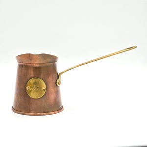 May include: A copper coffee pot with a long brass handle and a wide, flared rim. A circular brass plaque is attached to the front of the body, featuring the embossed number 50. The pot has a tapered cylindrical shape and a smooth metallic finish.