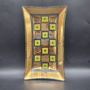 May include: Rectangular decorative glass tray with a gold border and a central checkered pattern. The squares alternate between brown, gold, and chartreuse green. The tray has a slightly curved shape and is likely used for display or serving.
