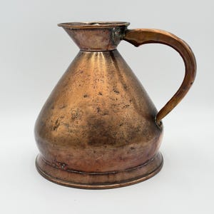 May include: Antique copper pitcher with a wide base and a narrow spout. The pitcher has a curved handle and a conical body. The surface shows signs of age and wear. The text "Gord. Craig's 1838" is visible on the body of the pitcher.
