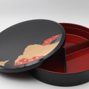 Pode incluir: Um prato de servir redondo preto dividido com interior vermelho e tampa. A tampa apresenta um design decorativo com detalhes bege, vermelhos e azuis. O prato está aberto, revelando os compartimentos internos.