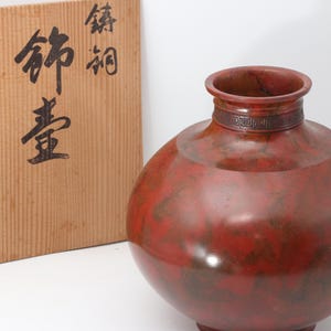 以下が含まれることがあります： 丸い胴体と細い首を持つ赤と茶色の装飾的な花瓶。 花瓶はテクスチャ加工された表面と首の周りに装飾的なディテールの帯があります。 日本語の文字が入った木箱が背景にあります。