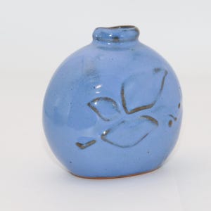 Puede incluir: Un pequeño jarrón redondo de cerámica azul con cuello estrecho. El jarrón presenta un diseño floral marrón y un acabado brillante. El jarrón mide aproximadamente 13 cm de alto y es adecuado para pequeños arreglos florales.
