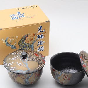 以下が含まれることがあります： 金、赤、白の草花と波の模様が描かれた、蓋付きの黒い陶器のボウル2つ。ボウルには、同じ草花の模様と「光琳絵図」などの日本語の文字が印刷された、黄褐色の段ボール箱が付属しています。