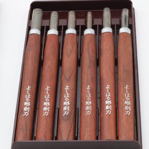 Op de afbeelding: Een set van zes houtsnijgereedschappen met bruine houten handvatten en zilveren bladen, netjes gerangschikt in een donkerbruine koffer. Elke handgreep heeft witte Japanse tekst. De gereedschappen zijn ontworpen voor gedetailleerd snijwerk.