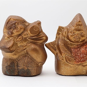 May include: Two ceramic Japanese figurines. The one on the left is a seated figure with a fish-shaped object. The one on the right is a figure holding a fish. Both are gold with brown and red accents.